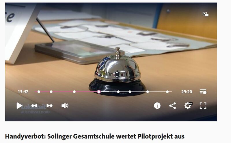 Auf einem Lehrerpult in einem Klassenraum liegen Unterrichtsmaterialien und eine Tischglocke Umgang mit Handys an der Gesamtschule Höhscheid