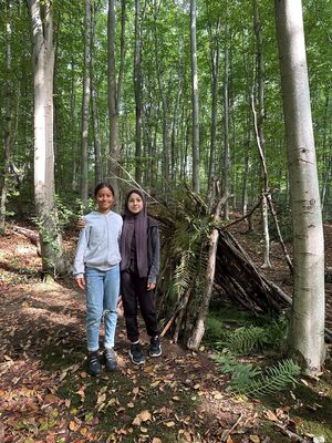 Zwei Schülerinnen präsentieren stolz ihr selbstgebautes Tipi aus Ästen und Farnen.