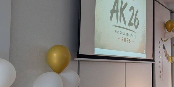 An der Präsentationsfläche ist eine Folie mit Abschlussklasse  (AK) 26 und dem Schullogo zu sehen.