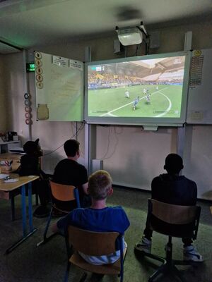 Vier Jungs sitzen in einem Klassenraum vor einer digitalen Tafel und zocken FIFA.