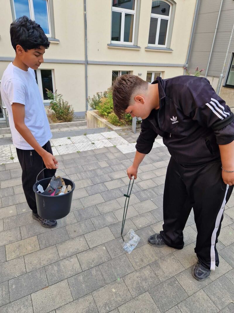 Zwei Jungs sammeln Müll mit einer Zange und einem Eimer auf dem Schulhof.