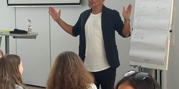 Herr Citir steht an einer Flipchart und bezieht sein Publikum mit ein. Im Vordergrund sind Schülerinnen und Schüler, die sich zu einem Redner im Publikum umdrehen.