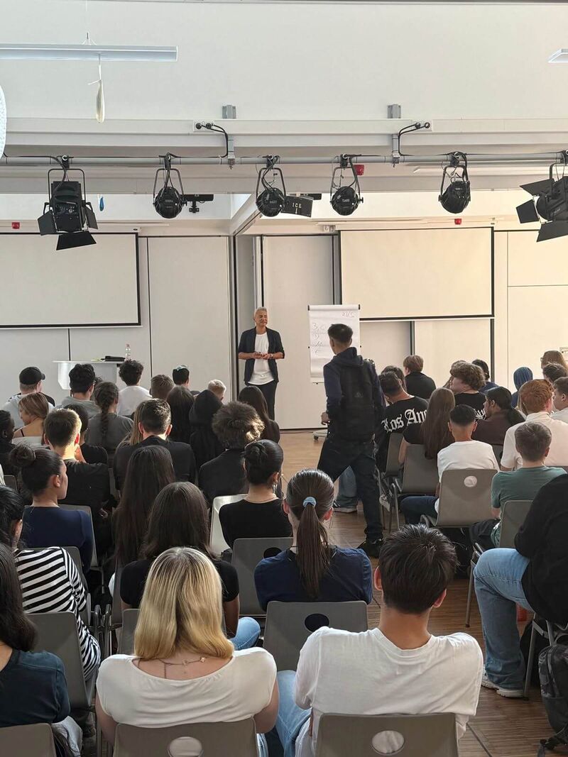 Herr Citir steht an einer Flipchart und bezieht sein Publikum mit ein, dass in Stuhlreihen sitzt. Ein Schüler kommt nach vorne.