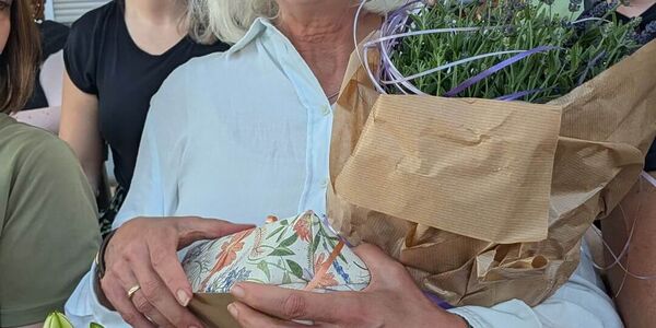 Eine strahlende blonde Frau hält Blumen im Arm und bekommt Geschenke.