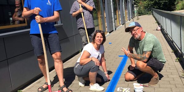 Eine Gruppe von Lehrkräften ist gerade dabei einen blauen Zahlenstrahl auf den Schulhof zu malen.