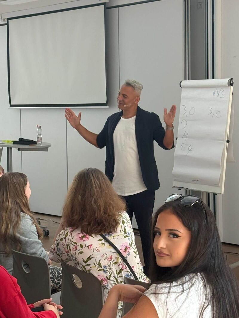 Herr Citir steht an einer Flipchart und bezieht sein Publikum mit ein. Im Vordergrund sind Schülerinnen und Schüler, die sich zu einem Redner im Publikum umdrehen.