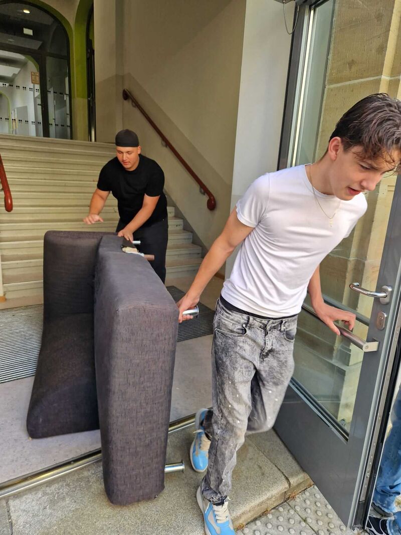 Zwei Jungs tragen ein altes Sofaelement durch den Haupteingang der Schule raus.