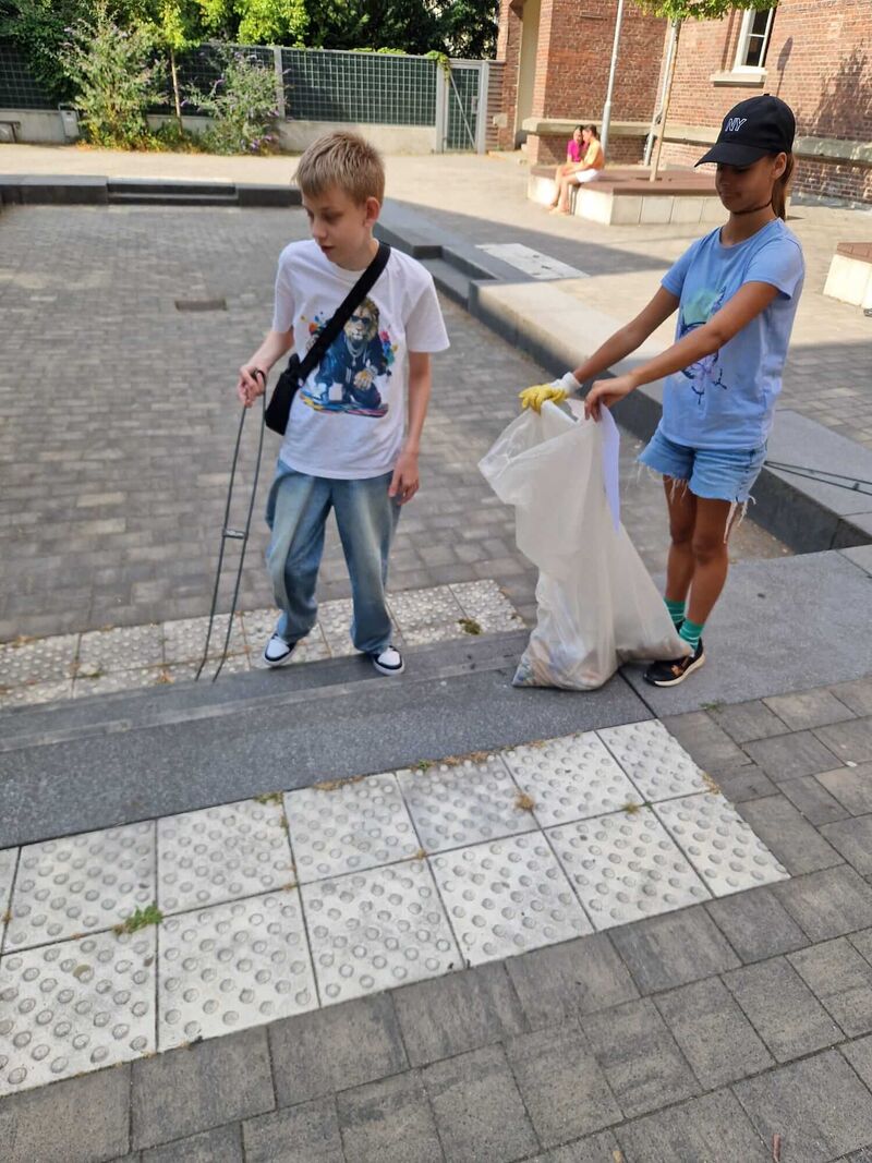 Zwei Kinder sammeln Müll mit einer Zange und einem Müllsack auf dem Schulhof.